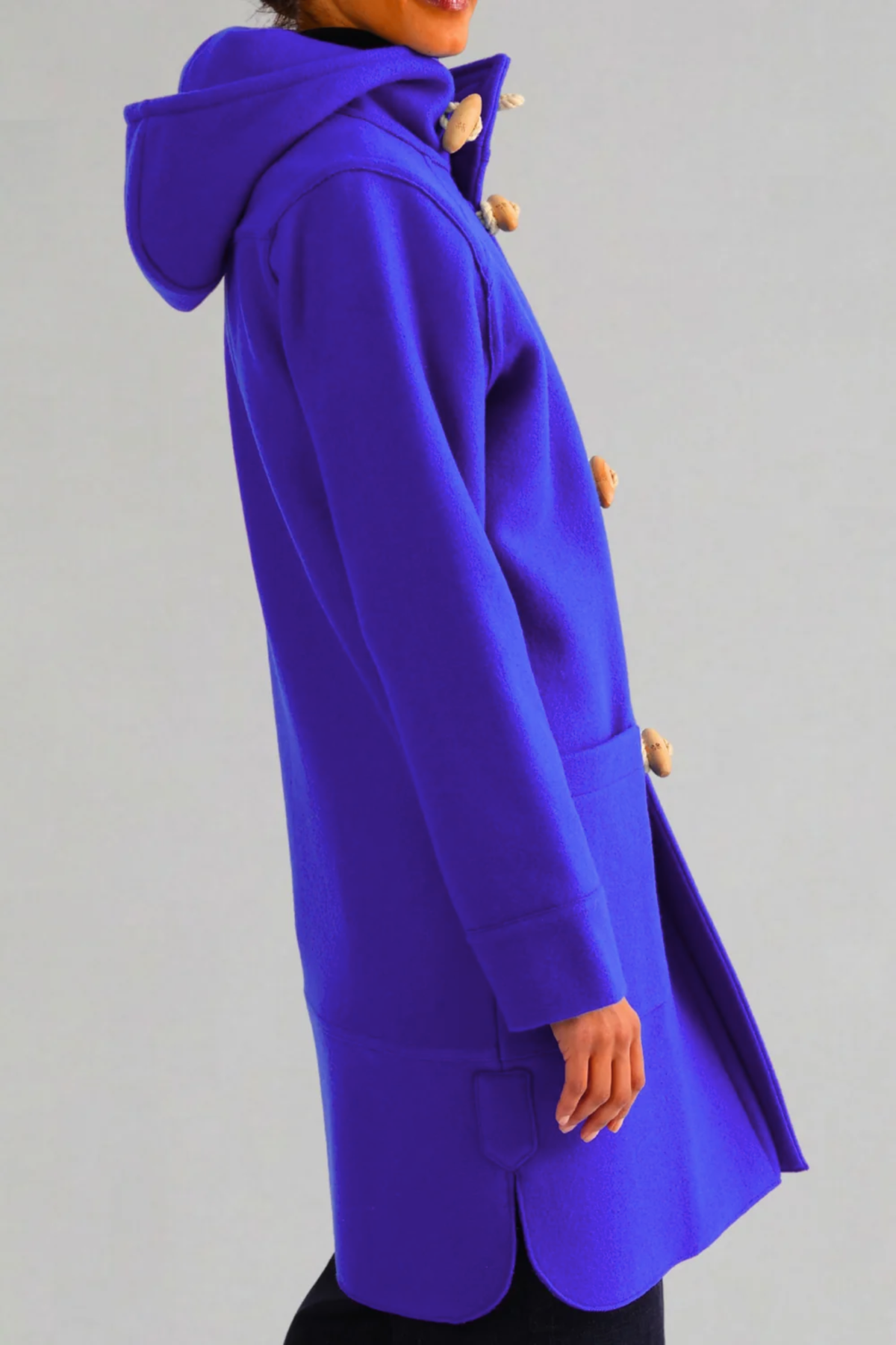 A person wearing a bright royal blue hooded duffle wool coat with tan toggle buttons, shown in side profile against a plain light gray studio background.