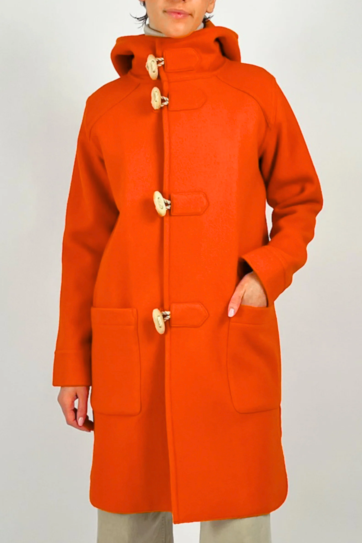 A person wearing a bright orange hooded duffel wool coat with large wooden toggle closures and front patch pockets, photographed against a plain light gray studio background.